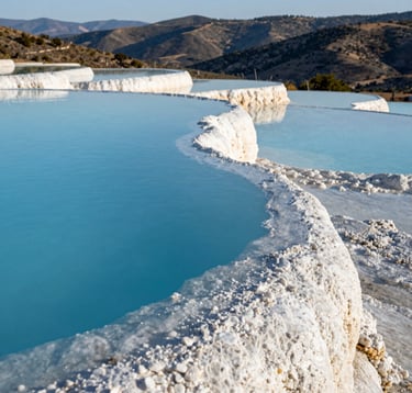 A close-up photograph of the natural mineral pools at Hierve el Agua. The water is a brilliant soft blue, reflecting the sky. The white, calcified edges of the pools create a stark contrast against the deep teal water. In the background, the rolling hills of Oaxaca, Mexico, are visible under a bright, clear sky.