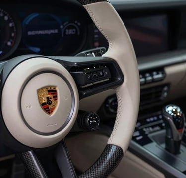 A close-up high-detail shot of a Porsche's steering wheel and gear shifter. The interior features soft off-white leather and carbon fiber accents. The lighting is moody, with deep night black shadows and soft midnight navy blue glows from the dashboard lights.