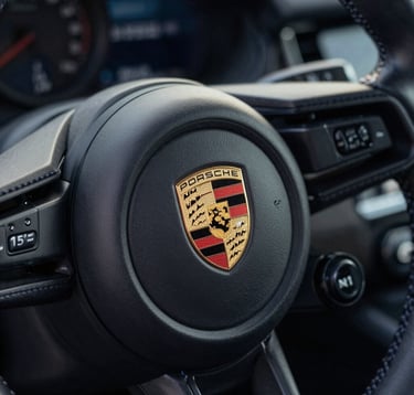 A detailed close-up shot of a Porsche steering wheel crest. The interior is Deep Obsidian Black leather with Midnight Navy contrast stitching, captured with a shallow depth of field in a Soft Pearl White light.
