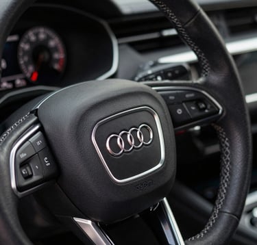 Close-up of a premium leather steering wheel in an Audi, with silver slate stitching and the brand logo. The dashboard in the background is dimly lit with pearl white indicators. Sophisticated texture detail.