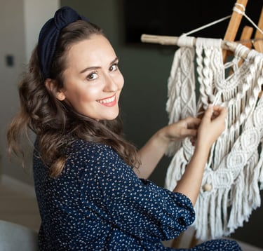 Mujer sonriente elaborando un tapiz de macramé blanco en un caballete de madera en casa.