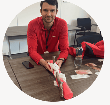 A person sits at a table holding a leg training model wrapped in a bandage, demonstrating first aid