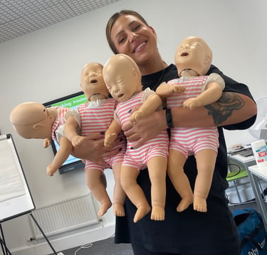A person smiles while holding four infant CPR training manikins in a classroom setting.