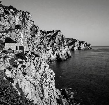 A high-contrast black and white photograph of a rugged Sicilian coastal landscape in Southern Europe, featuring dramatic cliffs and a small stone structure, capturing the somber and realistic mood of Verga's literary descriptions.