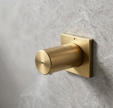 A close-up photograph of a sleek, minimalist steam outlet head made of brushed brass, integrated into a light grey marble shower wall. Delicate wisps of steam are emerging. The lighting is soft and natural, emphasizing the clean lines and high-end materials typical of a modern North American spa.