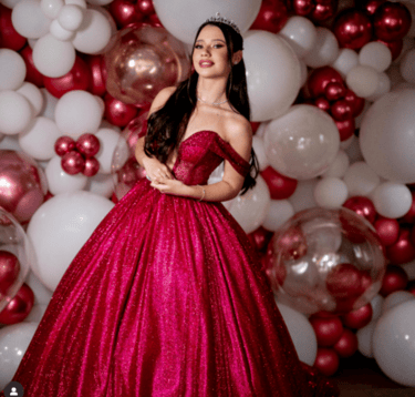 Debutante em vestido vermelho com tiara, em frente a painel de balões em cores vinho e branco, representando uma Festa de 15 