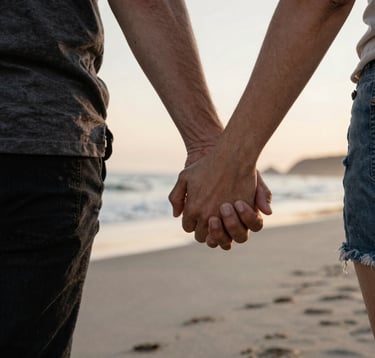 A candid, authentic photograph of a couple's hands interlaced while walking on a North American / US beach at sunset. The lighting is soft and sun-drenched. Hints of charcoal and warm brown appear in their clothing, contrasting softly with the sand and coastal water.
