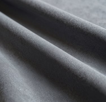 A close-up, high-fashion detail shot focusing on the texture of a luxury fabric. The color palette includes muted sky grey and deep charcoal tones. The lighting is soft and directional, emphasizing elegance and a refined artistic perspective.