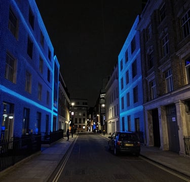 A high-resolution LiDAR laser scanning point cloud of a modern London street at night. The image consists of millions of vibrant electric blue data points forming the 3D outlines of buildings and a stationary vehicle against a deep charcoal void. Tech-forward, cybernetic aesthetic.