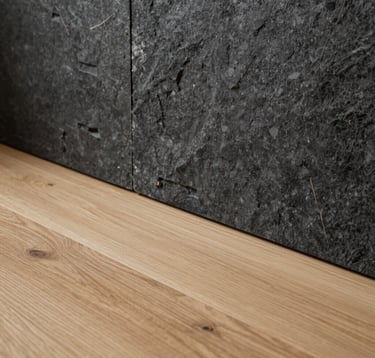 Close-up detail of high-quality renovation materials in a Northern European setting. A minimalist composition featuring light oak wood flooring meeting a dark graphite stone wall, with soft natural light highlighting the textures.