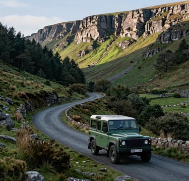 A classic green Land Rover driving along the winding Buttertubs Pass in the Yorkshire Dales. The road is flanked by steep dark forest green slopes and dramatic limestone cliffs. The lighting suggests a fresh, clear Northern European / British / Yorkshire morning.
