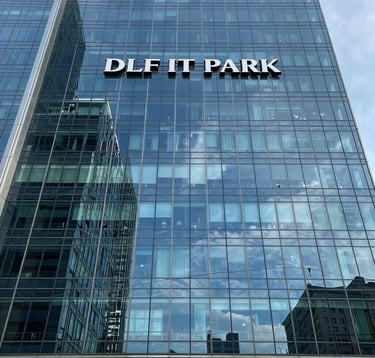 A professional architectural photograph of the DLF IT Park facade in Chennai. The building features sleek glass panels reflecting a steel blue sky, symbolizing modern commercial growth and high-value surroundings.