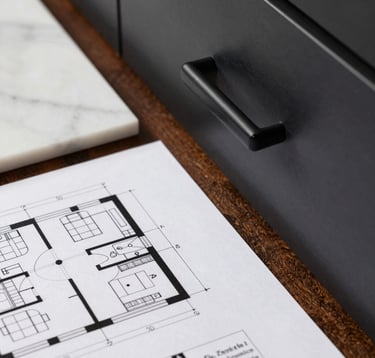 A detailed close-up of an architectural kitchen layout plan in a Los Angeles studio. Beside the plans lie a sample of white marble and a matte black cabinet handle. Professional lighting, precise and sophisticated photography.