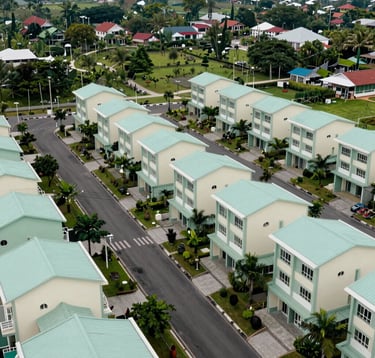 A wide photography shot of a well-planned residential community in a Southeast Asian / Indonesian valley. The image shows wide paved roads, underground utilities, and landscaped public parks. The composition is balanced and professional, featuring muted seafoam green and pale mint cream colors in the architecture.