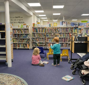 Toddlers exploring books in the children’s area of Saffron Walden Library, low shelves, colourful chairs & space for buggies.