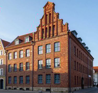 A professional architectural photograph of the historical brick administrative buildings at Gdansk Seaport. The red bricks are illuminated by a soft white morning sun, with dusty blue window frames and a clean, steel blue sky in the background.