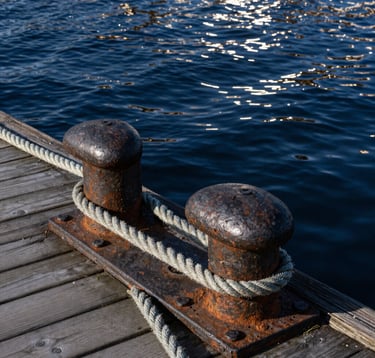A close-up photograph of weathered iron bollards and thick ropes on a wooden pier in Gdansk. The water in the background is a deep Midnight Navy Blue, and the sunlight reflects a Soft Frost White glint off the ripples.