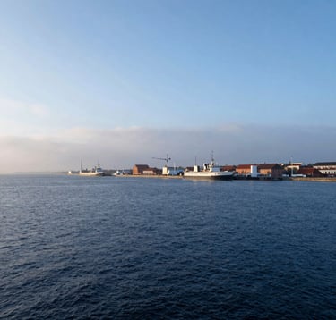 A serene photograph of the Gdansk coastline where the port meets the open sea. The water is a deep navy blue, contrasting with the soft misty blue and cloud white of the horizon. Elegant and inspiring perspective.