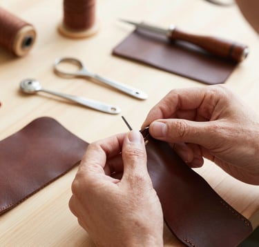 A detailed shot of an artisan's hands meticulously stitching a piece of high-quality dark brown leather. The setting is a bright, minimal workshop in Western Europe with silver tools arranged neatly on a wooden workbench. The mood is focused and serene, emphasizing premium quality and heritage craftsmanship.