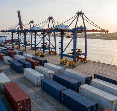 A high-perspective photograph of a modern industrial commodity port at sunrise. Rows of shipping containers and large-scale infrastructure. Professional, authoritative, clean composition. Global / Emerging Markets. Colors include dark blue and off-white.