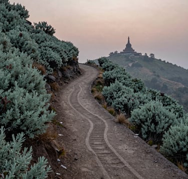 An elegant photo of a winding tracking route carved into the side of Phulchoki hill. The path is surrounded by sage green shrubs, with a distant, misty view of a traditional stupa silhouette against the horizon at dusk.