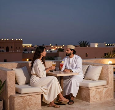 An elegant North African / Moroccan couple enjoying cocktails at dusk on a modern, minimalist rooftop bar. The design features Soft Sand stone and Ivory White cushions. In the background, the soft lights of Marrakech begin to twinkle under a deep Indigo sky.