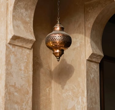 A close-up of an elegant interior in a North African / Moroccan riad. The walls are finished in smooth tadelakt of warm beige, and a single artisanal copper lamp in muted gold hangs from the ceiling. Dark charcoal shadows create a sense of depth and exclusive mystery. Sharp focus on textures.