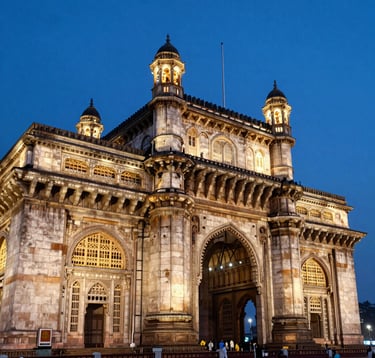 A vibrant high-resolution photograph of the illuminated Gateway of India in Mumbai at dusk. The architectural detail is sharp against a deep blue evening sky, with professional lighting creating a warm off-white glow on the stone.