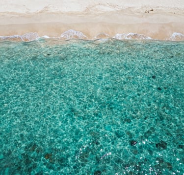 An aerial top-down photograph of a remote, crystalline turquoise lagoon in the Indian Ocean, with a thin strip of ivory sand. The composition is minimalist and spacious, capturing the essence of private access and solitude for the Global / Discerning English-speaking elite.