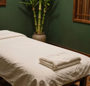 A close-up shot of a serene Indonesian massage room in South Jakarta. A massage table with pristine off-white linen, green bamboo shoots standing in a ceramic vase nearby, and warm soft lighting casting a gentle glow on dark forest green walls. Natural wood textures, peaceful luxury spa mood.