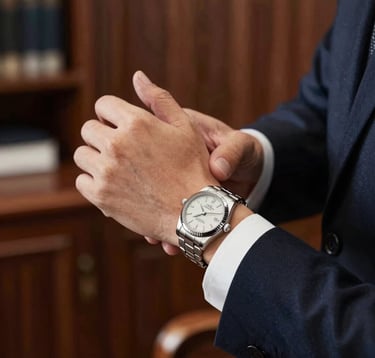 Close-up of a man's hands adjusting a luxury silver wristwatch (#7D8C9C). He is wearing a crisp white shirt and a dark navy suit sleeve. The background is a blurred high-end executive office with mahogany textures. Lighting is soft and professional.