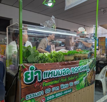 Street food vendor in Thailand serving fermented pork salad with fresh greens and vegetables at a night market.