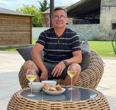 A man wearing glasses relaxes in a wicker chair outdoors with white wine and appetizers.