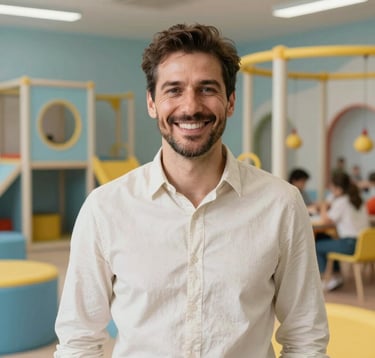 Portrait of a friendly man in his early 40s with a warm smile, wearing a casual shirt in Off-white. He is standing in a brightly lit, modern children's activity center in Southeast Europe. The background features safe, colorful play equipment in Light Blue and Yellow.