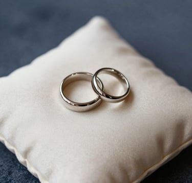 A macro photograph focusing on the fine details of a luxury wedding: a pair of bespoke rings resting on a soft cream silk cushion. The lighting is soft and ethereal. Background includes hints of deep navy blue and slate gray-blue textures. Sophisticated and minimalist composition, Hispanic setting context.