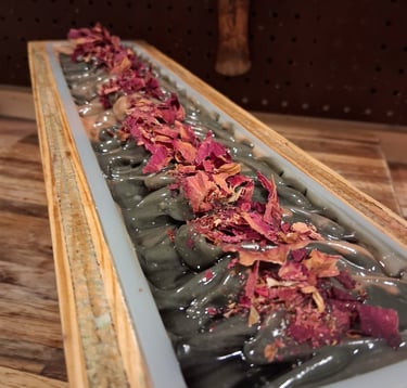 Handmade artisan loaf of dark charcoal soap topped with dried rose petals in a wooden mold.
