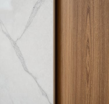 A close-up artistic photograph of premium architectural finishes: white marble with grey veins meeting a polished oak wood panel. Soft, natural side-lighting emphasizes the textures and the minimalist, solid construction. Spanish-speaking / Iberian Peninsula.