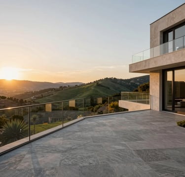 Wide-angle photography of a luxury estate's private terrace overlooking a valley in the Spanish-speaking / Iberian Peninsula. The foreground shows a minimalist grey stone floor and a glass railing. The sun is setting, casting a warm golden glow over the green hills and the sophisticated architectural lines of the building.