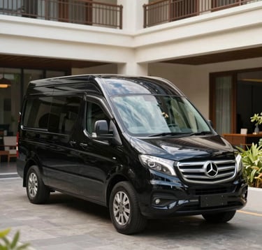 A luxury black transport van parked in front of a modern Bali hotel. The vehicle is clean and reflective, signifying professional reliability. The lighting is bright day light, emphasizing the #1B2D3B sleekness of the car against a #FDF6ED architectural background.