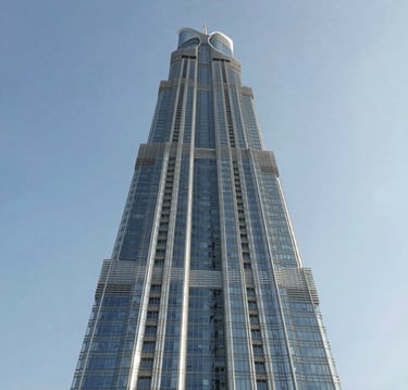 An upward architectural photography shot of a tall, modern skyscraper in a Middle Eastern urban setting. The structure uses reflective glass and silver metal frames, with a clear blue sky in the background. Minimalist and imposing.