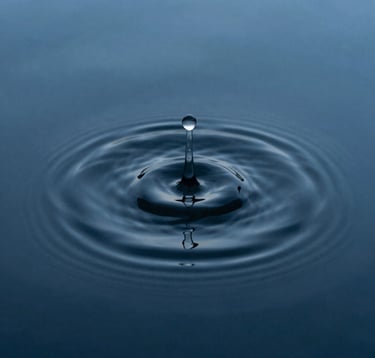 A minimalist macro photograph of a single ripple in a dark, steel blue pool of water. The composition is centered and calm, emphasizing perfect symmetry and the beauty of a single movement in a quiet landscape.