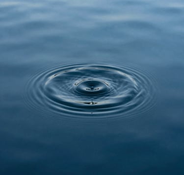 A minimalist photograph of a single ripple expanding on the surface of a deep steel blue pool of water. The composition is tight and focused, emphasizing the profound stillness of the surrounding liquid. Soft, diffuse lighting.