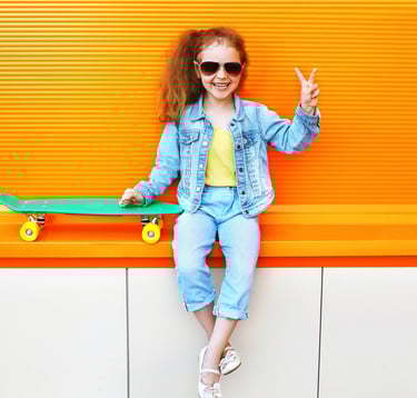 Little girl with a skateboard and holding up a peace sign, wearing kid's athletic sunglasses
