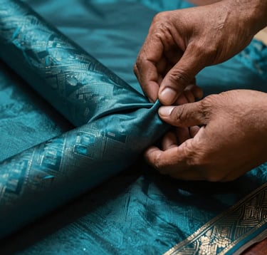 A close-up photograph of a South Asian / Indian artisan's hands carefully inspecting a roll of premium deep teal silk fabric. The lighting is focused and artistic, with metallic silver highlights on the texture of the cloth, emphasizing luxury and quality control.