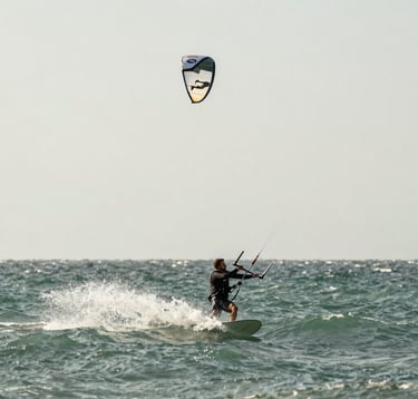 A dynamic action shot of a kitesurfer catching the wind, captured with high-speed shutter for crisp water droplets. The lighting is brilliant and natural. The ocean is a deep sage green #7E8A7F, and the sky is a pale off-white #FAF8F5. Minimalist composition.