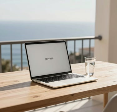 A sophisticated, minimalist workspace on a balcony overlooking the Mediterranean. A premium laptop and a single glass of water sit on a light wood desk. The aesthetic is clean, intellectual, and wealthy, using the brand's palette of sand and cream.