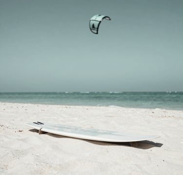A minimalist, high-end photograph of a kite surfing board resting on pristine white sand. The board features clean lines and a subtle matte finish. In the background, the soft gradient of a sage-colored ocean meets the sky. Aspirational and calm luxury aesthetic.