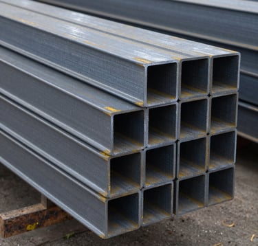 A clean, organized stack of heavy structural steel I-beams in a North American warehouse. The lighting is crisp and cool, highlighting the metallic texture and precision of the fabrication. Shot from a medium distance at eye level.