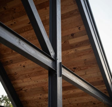 Close-up detail of the modern A-frame structure showing the intersection of charcoal black steel beams and warm wooden cladding. Sophisticated architectural textures under soft sunlight.