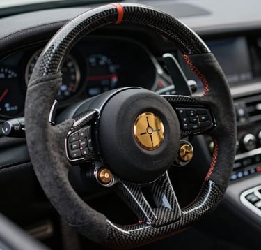 A close-up photograph of a professional racing steering wheel in a luxury car. The texture of carbon fiber and Alcantara is visible. The lighting is focused and dramatic, highlighting the buttons and dials in muted metallic gold against charcoal grey shadows.